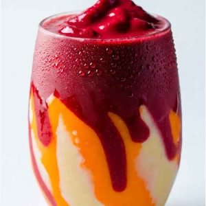 Thick mixed fruit smoothie with berries, bananas, and mango in a clear glass with condensation on a bright background.